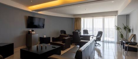 A modern hotel lobby with a long sofa, a coffee table, a TV mounted on the wall, and a large window with curtains.