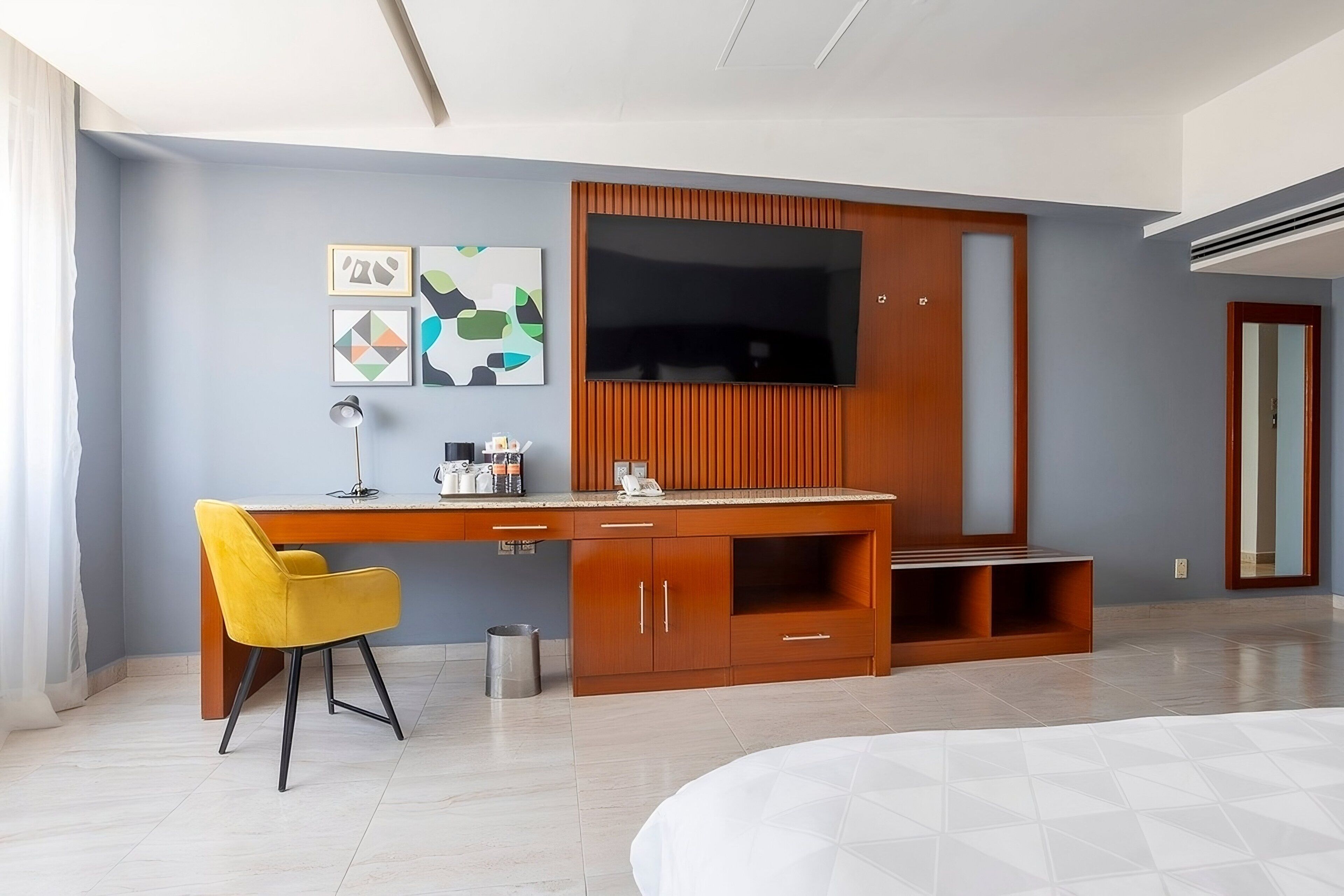 Una habitación de hotel moderna con un escritorio de madera, una silla amarilla, un televisor de pantalla plana y arte abstracto en la pared.