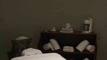 A hotel room with a bed, a bedside table with a coffee maker, and a shelf with towels.