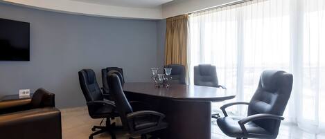 Uma sala de conferências com uma mesa de madeira escura, quatro cadeiras de escritório pretas, uma grande televisão de tela plana na parede e uma janela grande com cortinas finas.