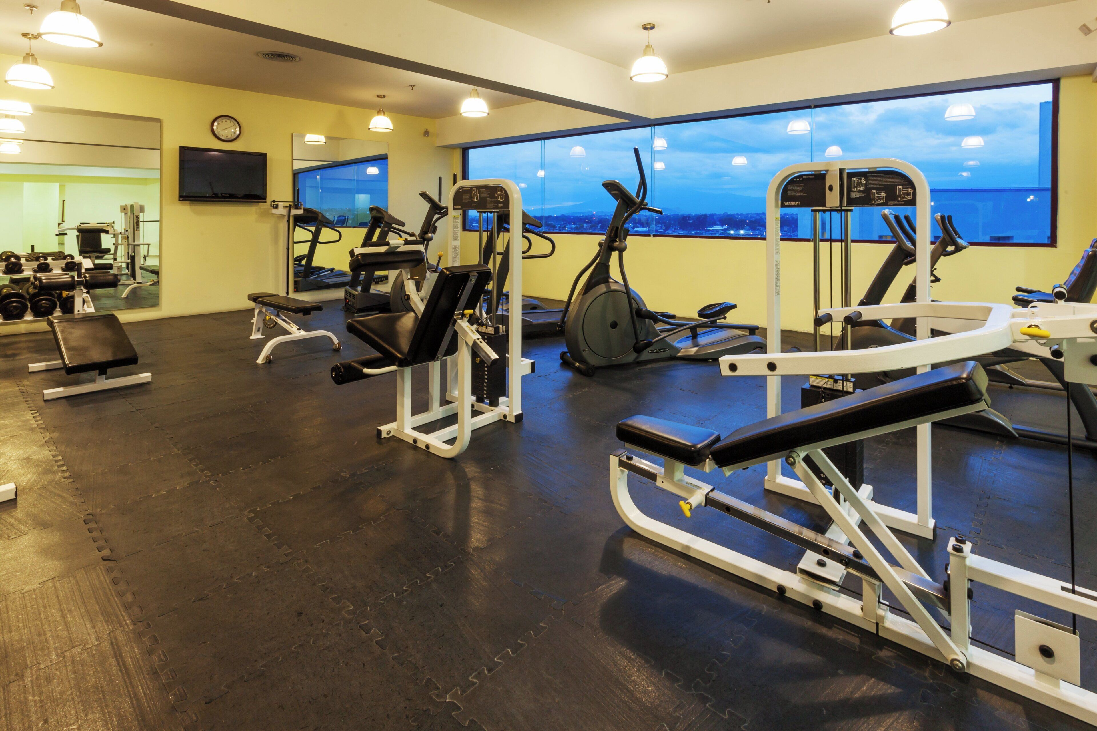 Ein Fitnessraum mit unterschiedlichen Trainingsgeräten, einem großen Fenster mit Blick und einer Uhr an der Wand.
