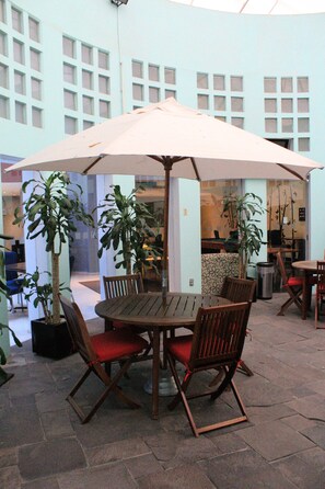 An outdoor patio area with a round wooden table and chairs, a large umbrella, potted plants, and a tiled floor. - Holiday Inn Puebla Finsa by IHG (Puebla)