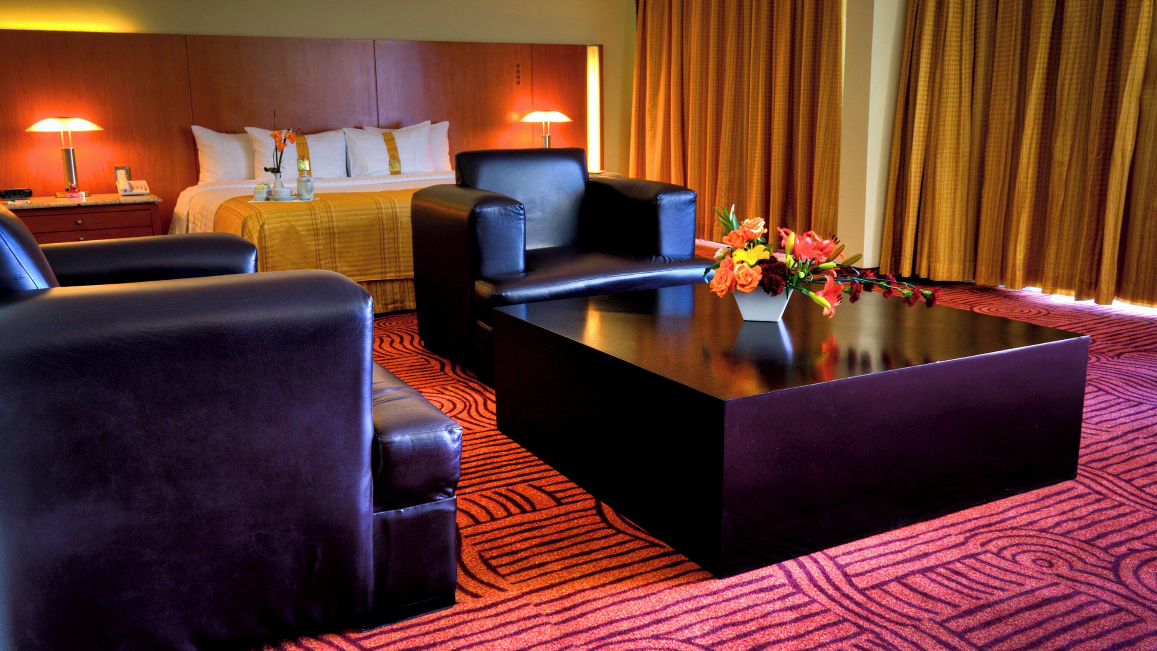 A hotel room with a bed, two armchairs, a coffee table with a vase of flowers, and a nightstand with a lamp.