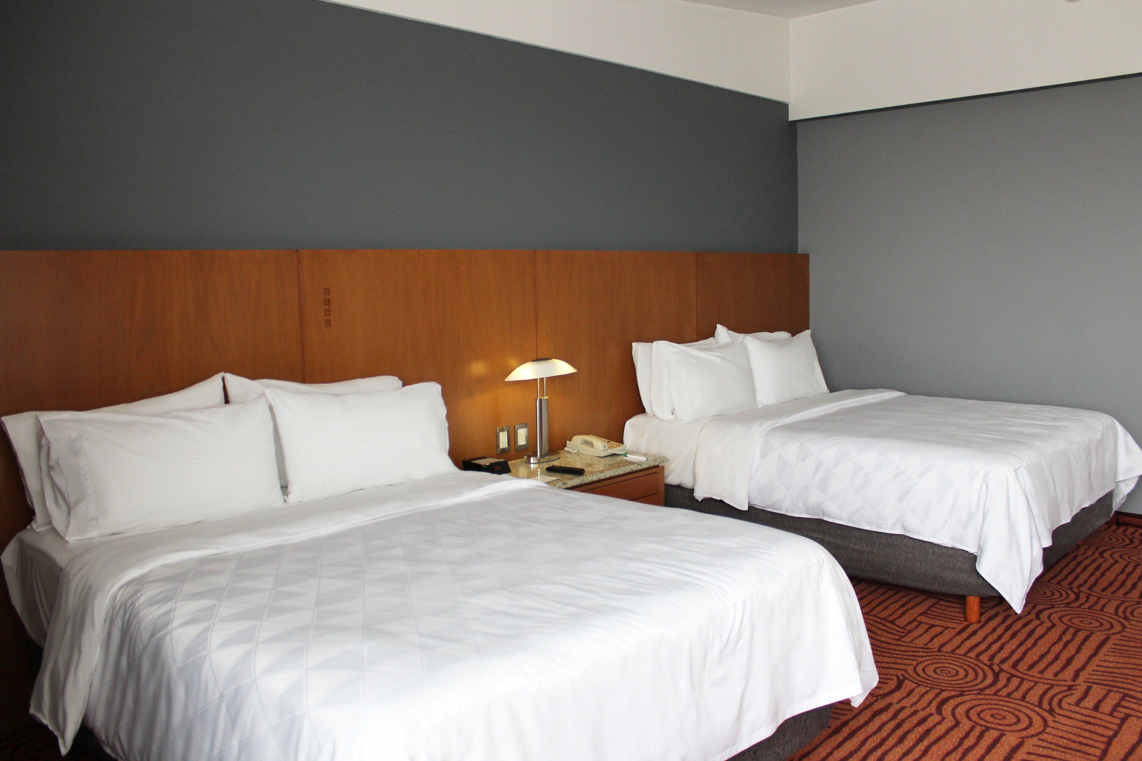 A hotel room with two beds, a nightstand with a lamp, and a carpeted floor.