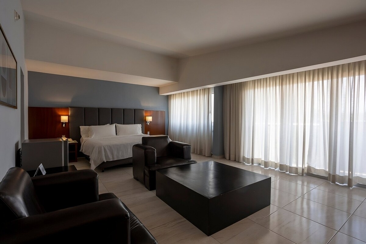 A modern hotel room with a bed, a desk, a coffee table, and a large window with curtains.