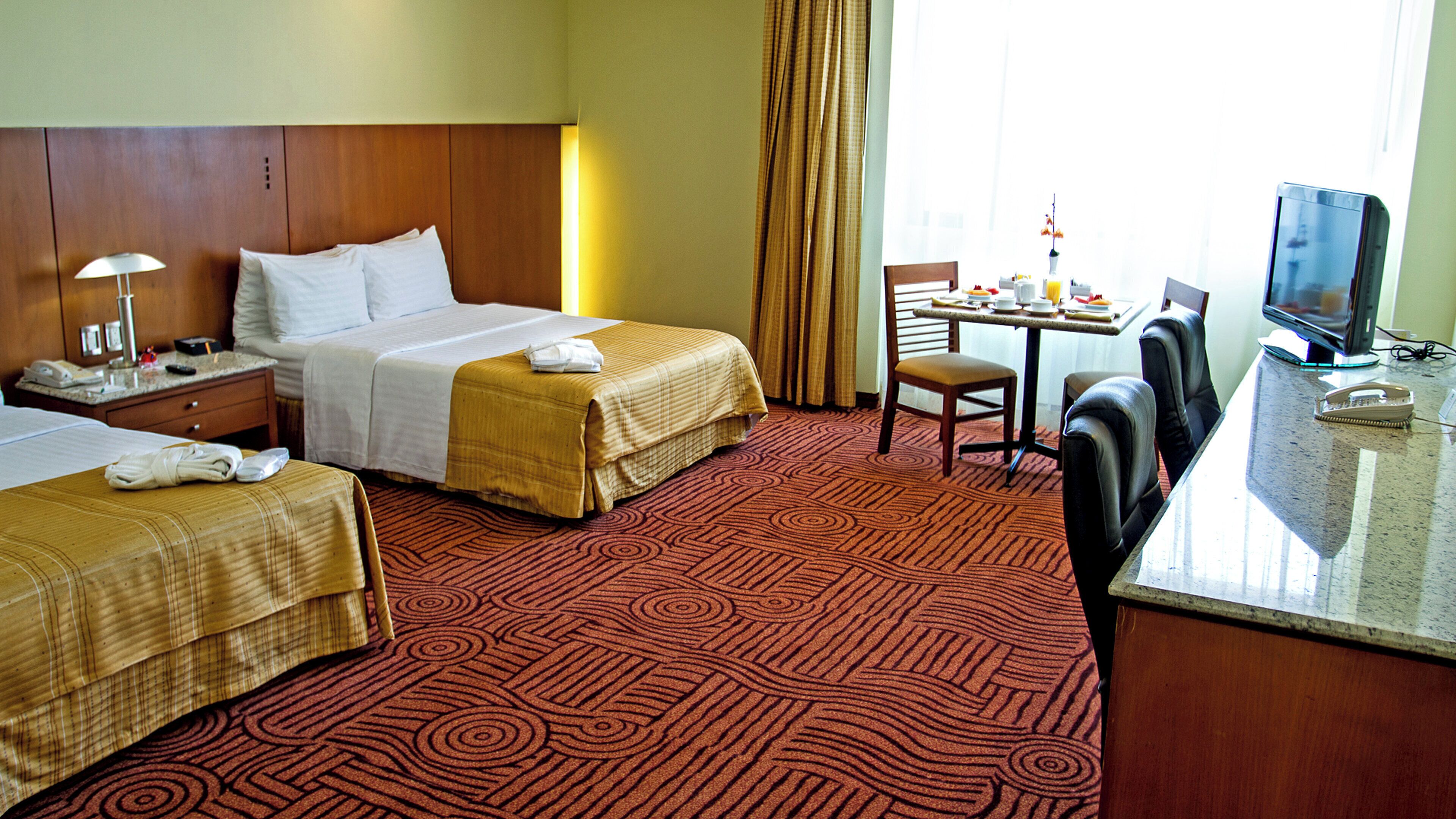 A hotel room with two beds, a desk, a chair, a television, and a table with breakfast items.