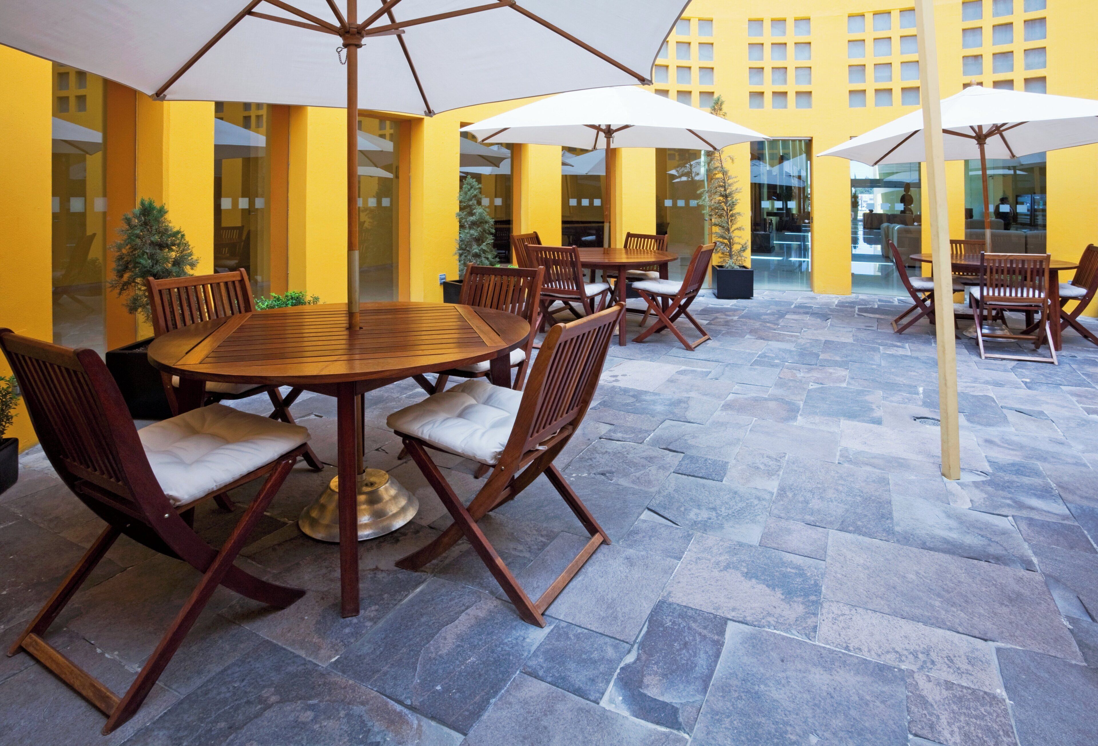 A patio area with round wooden tables and chairs, surrounded by yellow walls and large windows.