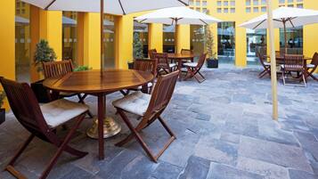 A patio area with round wooden tables and chairs, surrounded by yellow walls and large windows.
