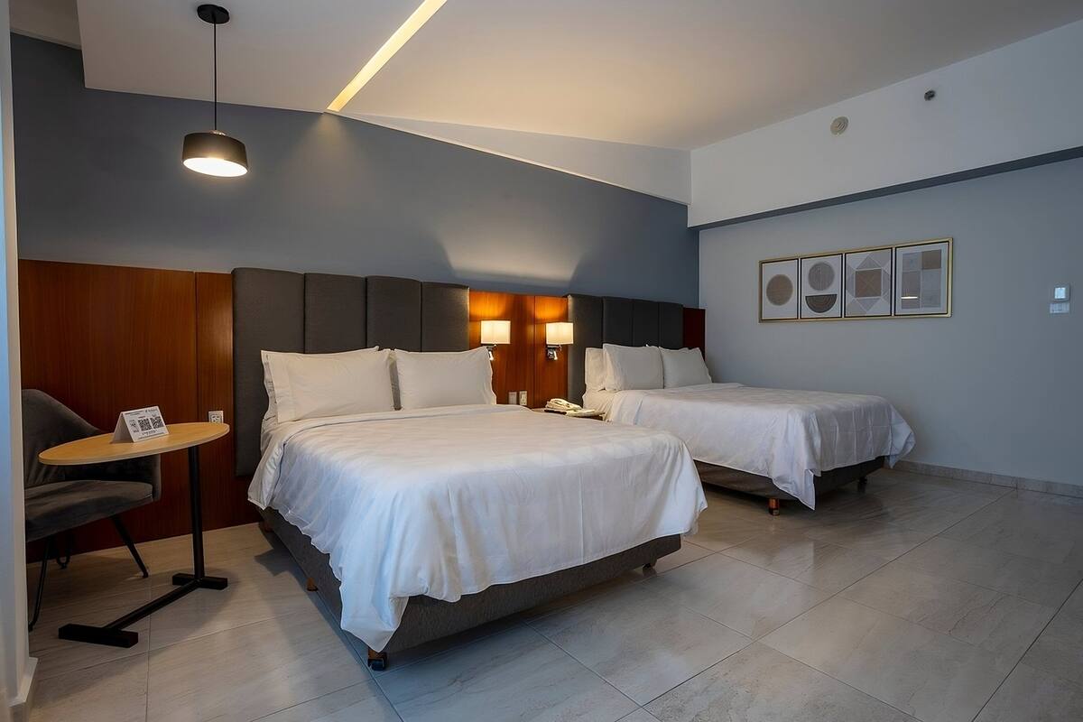 A hotel room with two beds, a small table, and a chair. There is a wall-mounted lamp and a framed picture above the beds.