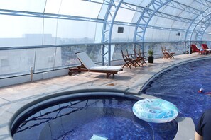 An indoor swimming pool with clear glass walls, lounge chairs, and a floating ring.