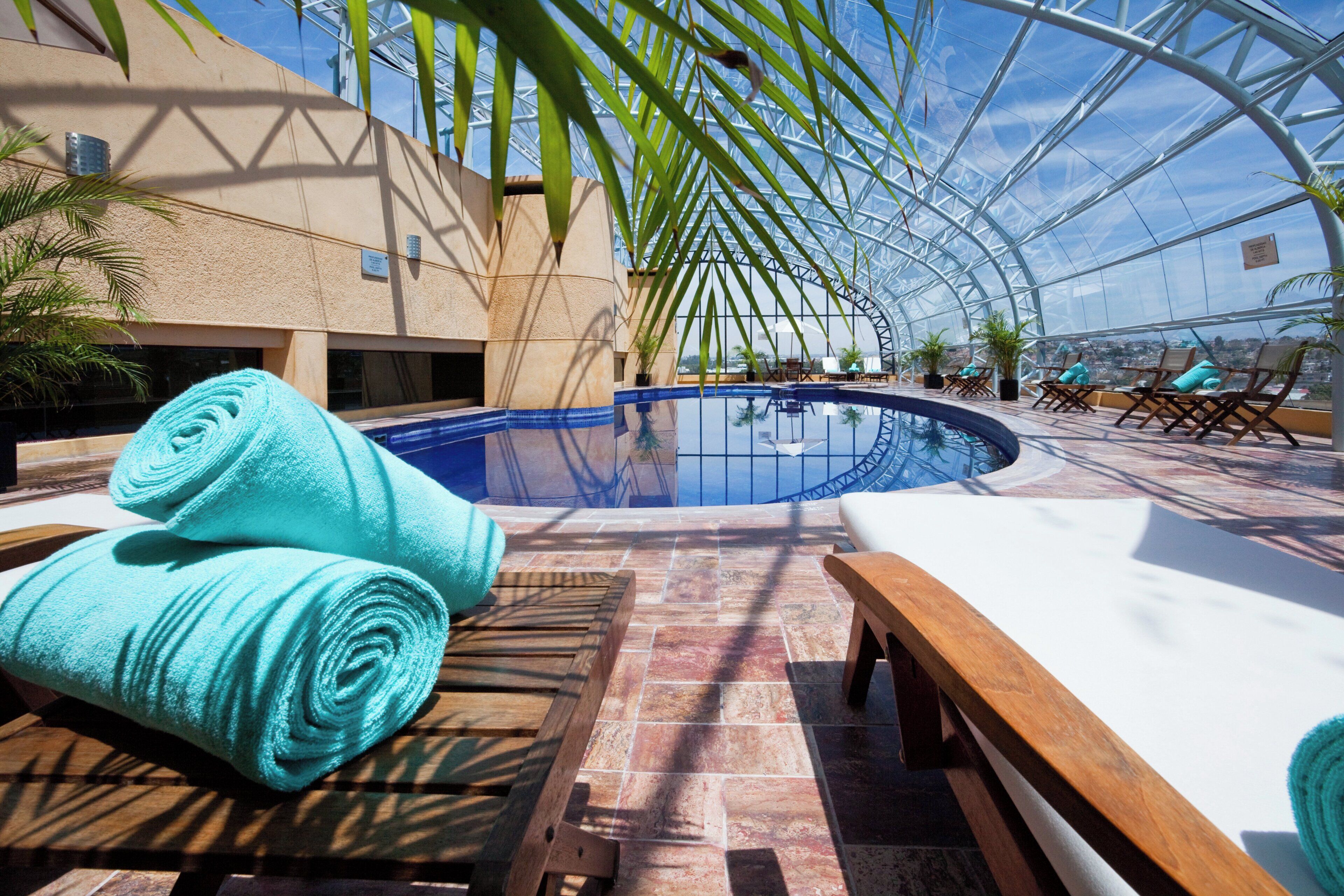 A rooftop pool area with a clear view of the city, a pool, lounge chairs, and towels.