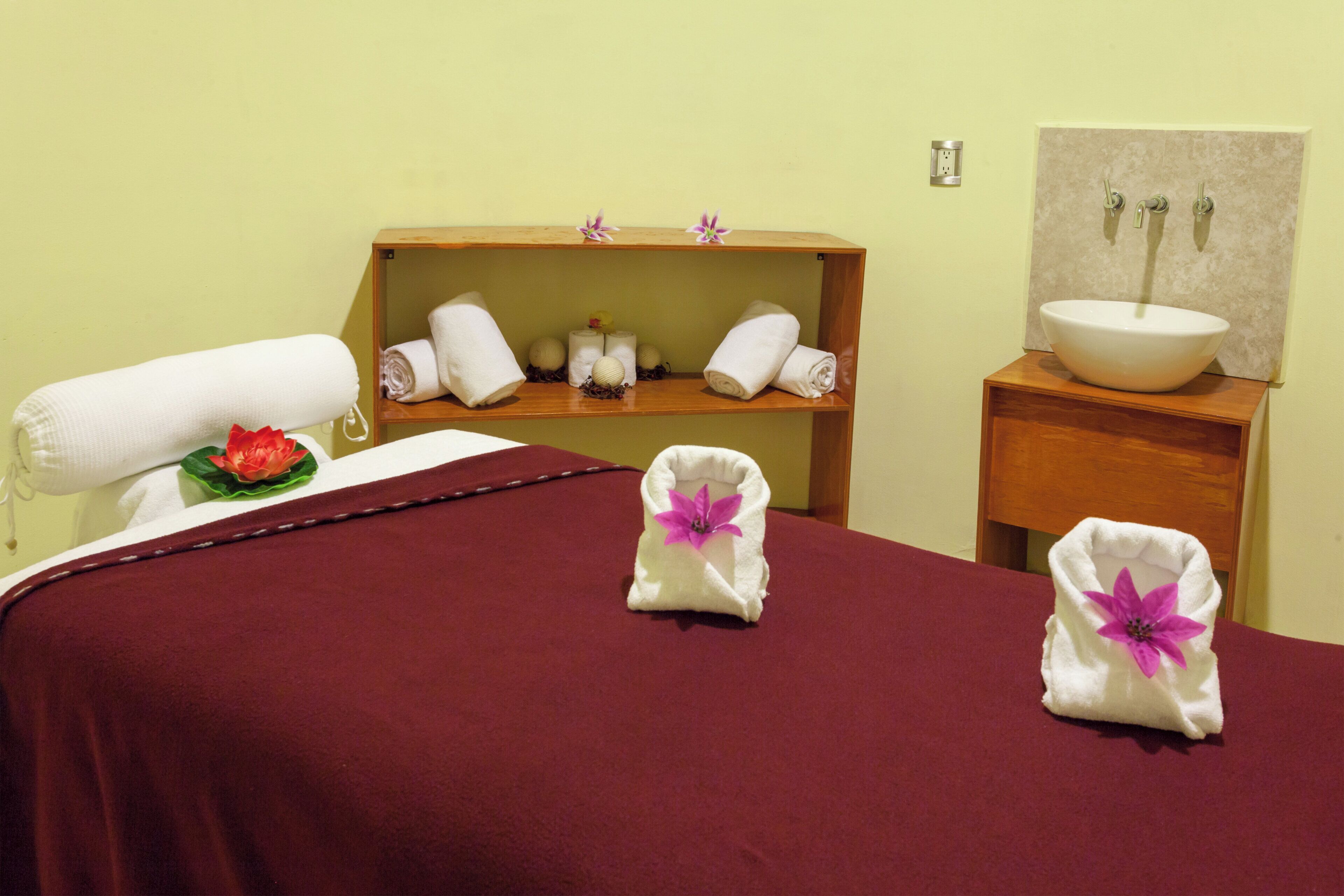 A hotel room with a bed, a wooden shelf with towels and toiletries, a wooden nightstand with a bowl sink, and a wall-mounted towel rack.