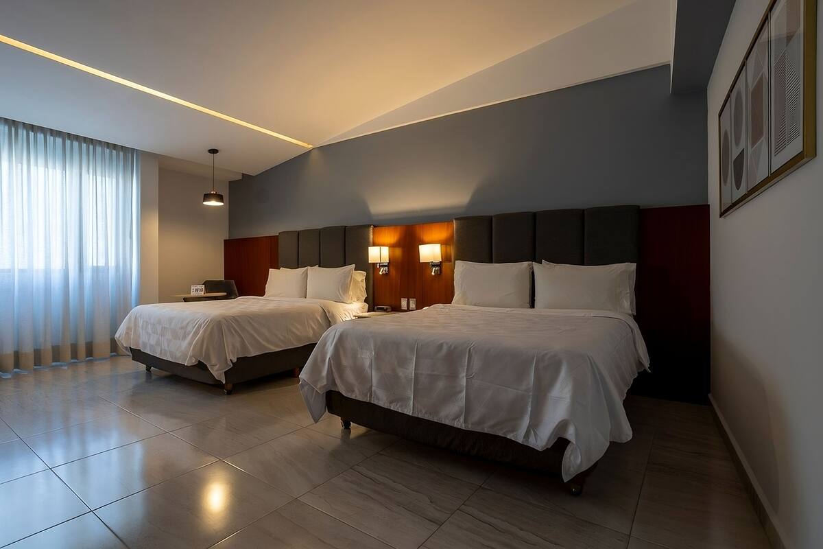 A hotel room with two beds, a large window with curtains, a modern headboard, and a wall-mounted light fixture.