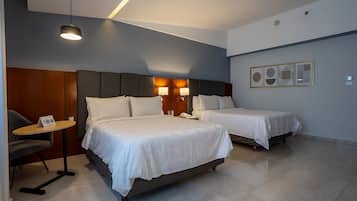 A hotel room with two beds, a small table, and a chair. There is a wall-mounted lamp and a framed picture above the beds.