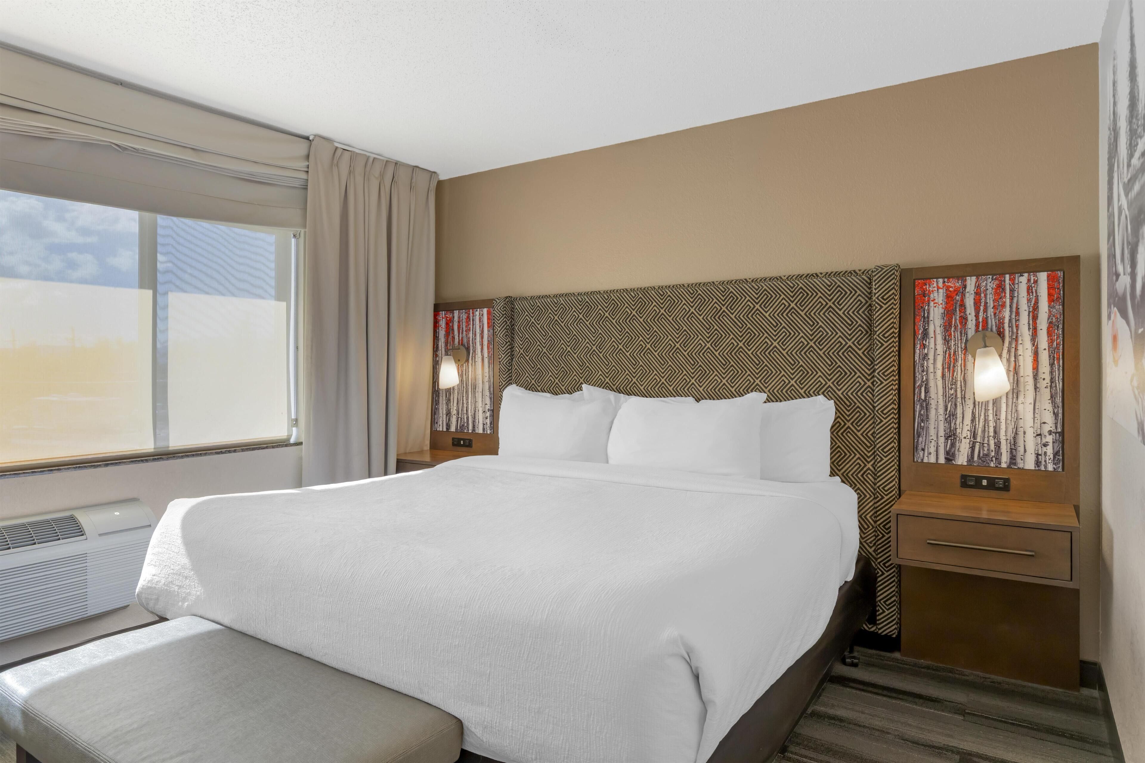 Una habitación de hotel con una cama grande, una mesita de noche con lámpara y una ventana con cortinas.