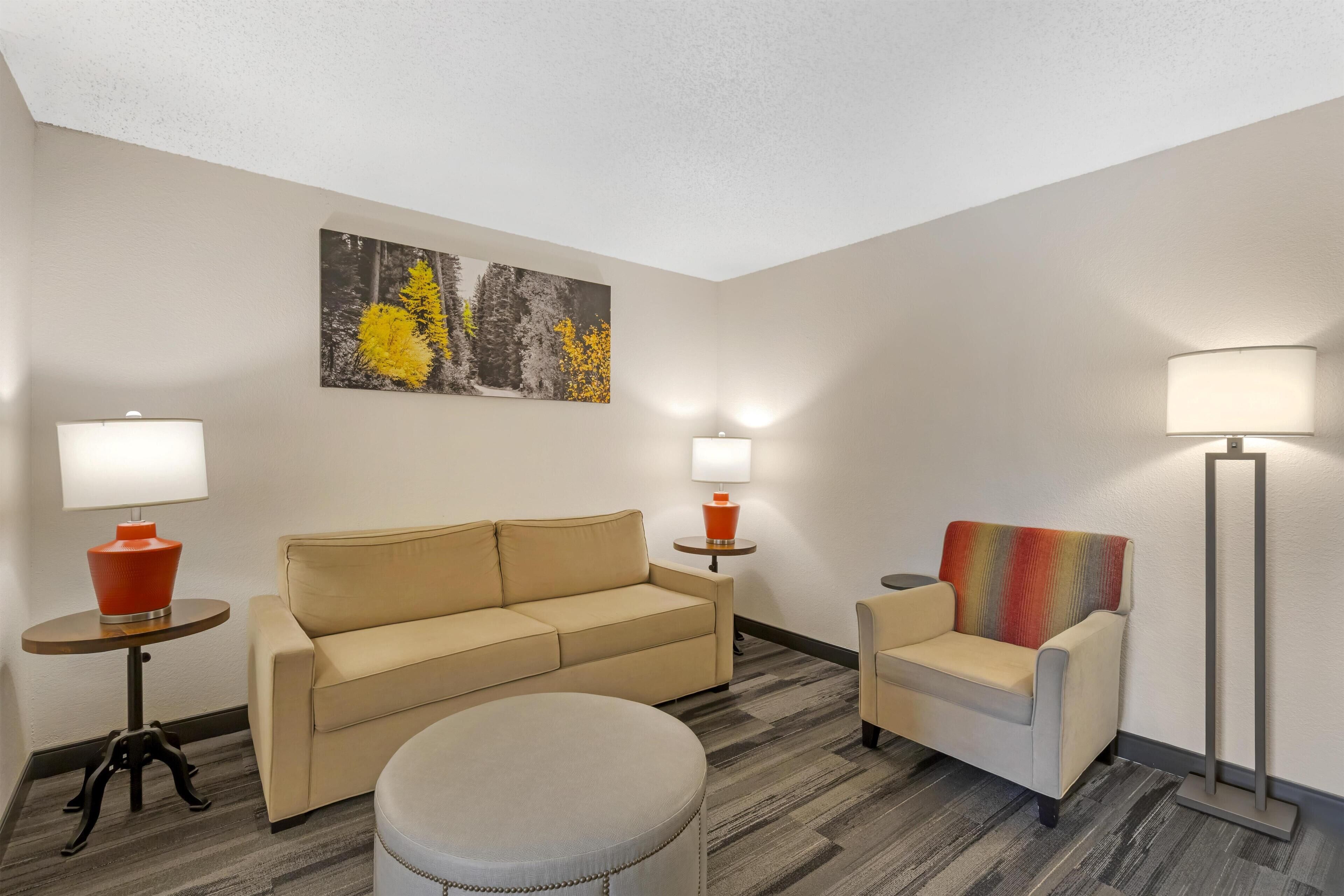 A hotel room with a beige sofa, a small round ottoman, a colorful armchair, two side tables with lamps, and a large abstract painting on the wall.