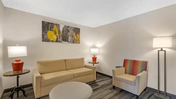 A hotel room with a beige sofa, a small round ottoman, a colorful armchair, two side tables with lamps, and a large abstract painting on the wall.