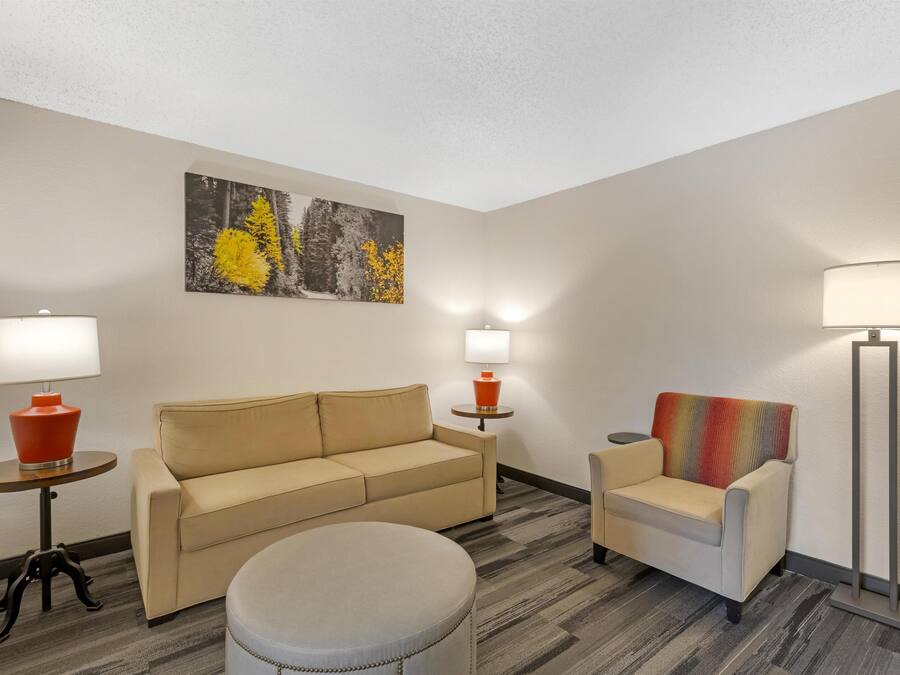 A hotel room with a beige sofa, a small round ottoman, a colorful armchair, two side tables with lamps, and a large abstract painting on the wall.