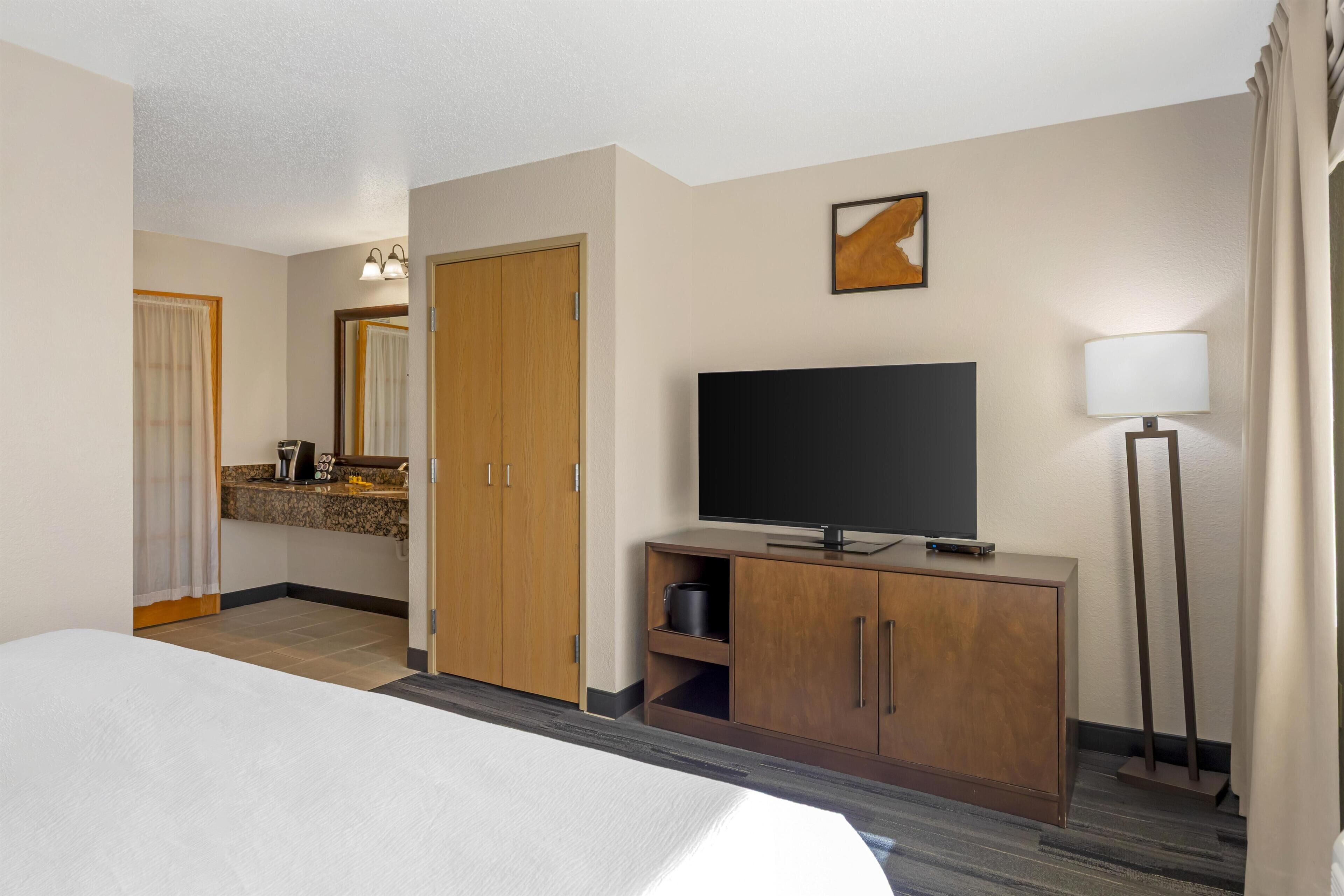 Habitación de hotel con cama, televisión sobre un mueble de madera, escritorio con lámpara y baño con cortina de ducha.