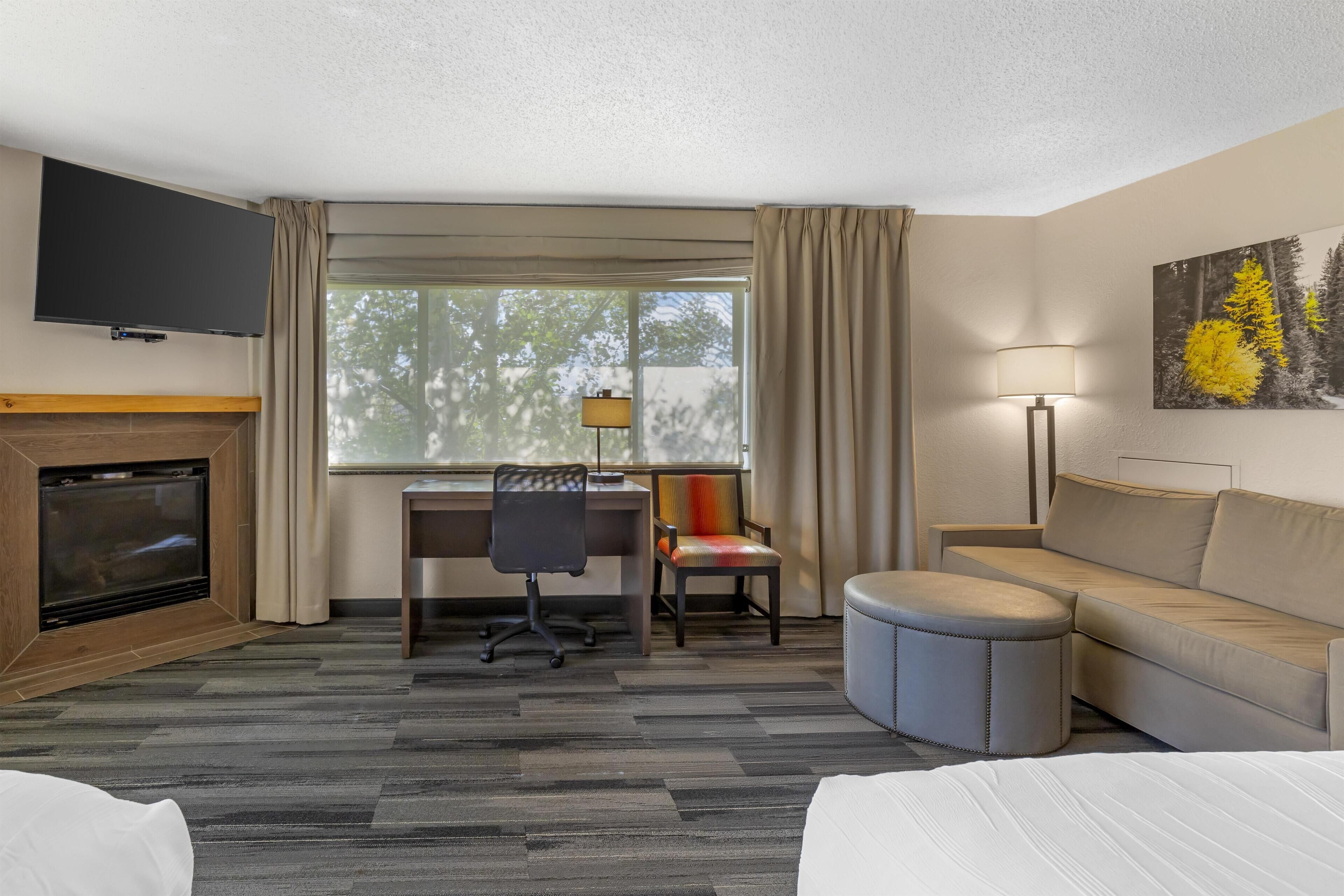 A hotel room with a TV, fireplace, desk, chair, couch, ottoman, and a painting on the wall.