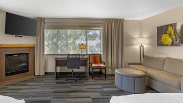 A hotel room with a TV, fireplace, desk, chair, couch, ottoman, and a painting on the wall.