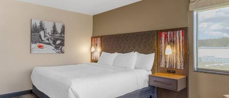 Een hotelkamer met een groot bed, een nachtkastje met lamp, een hoofdeinde met een patroon en een raam met uitzicht, plus een kunstwerk aan de muur.