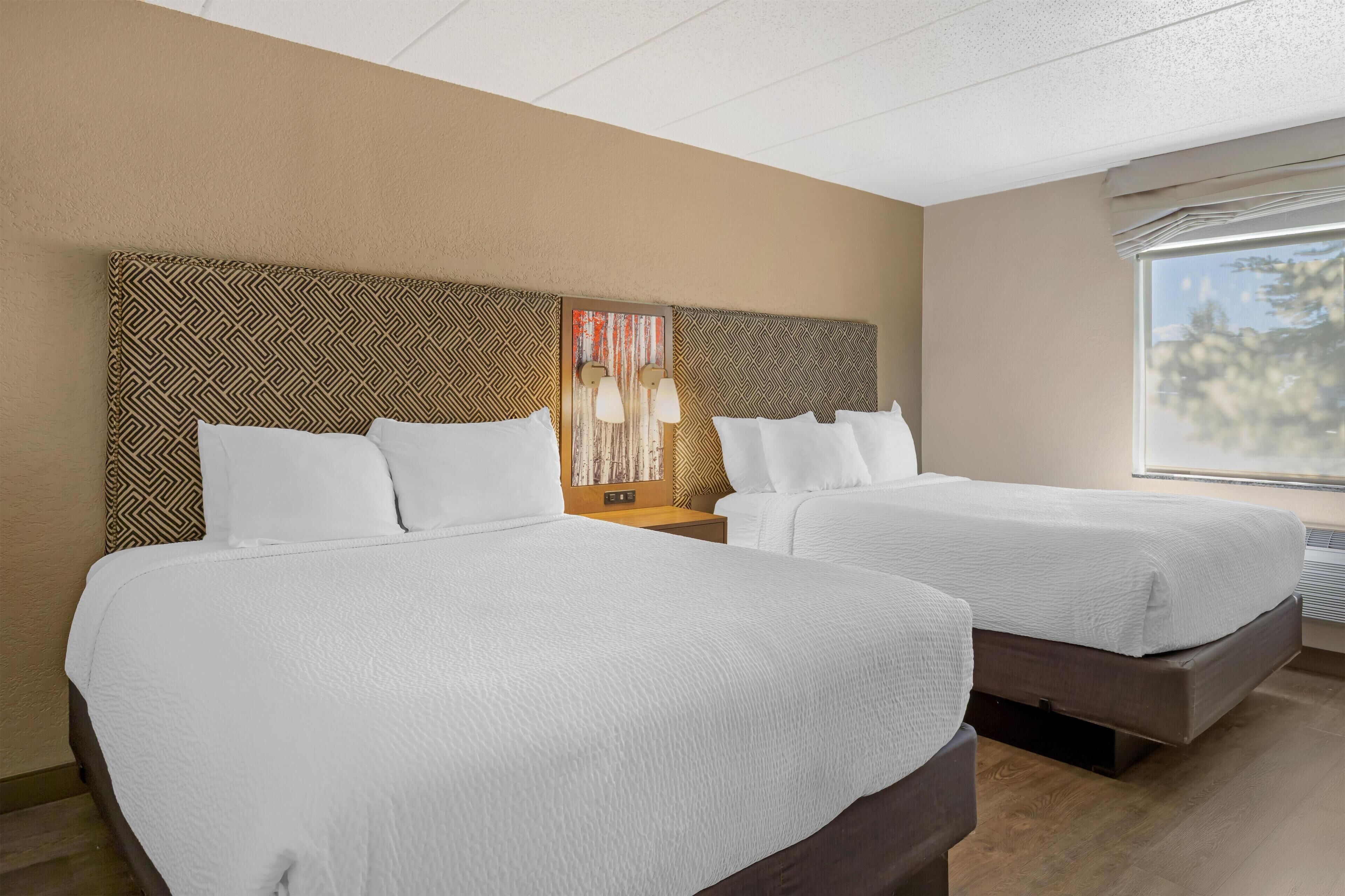 A hotel room with two beds, a patterned headboard, a wall art piece, and a window with a view of trees.