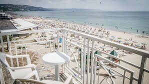 A beach view from a balcony with white chairs and a small table, overlooking a crowded beach with umbrellas and people.