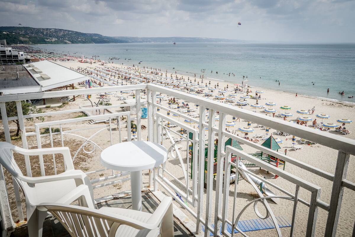 A beach view from a balcony with white chairs and a small table, overlooking a crowded beach with umbrellas and people.