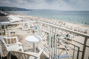 A beach view from a balcony with white chairs and a small table, overlooking a crowded beach with umbrellas and people.