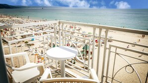 Un balcón con sillas y una mesa blancos, con vistas a una playa con sombrillas y gente.