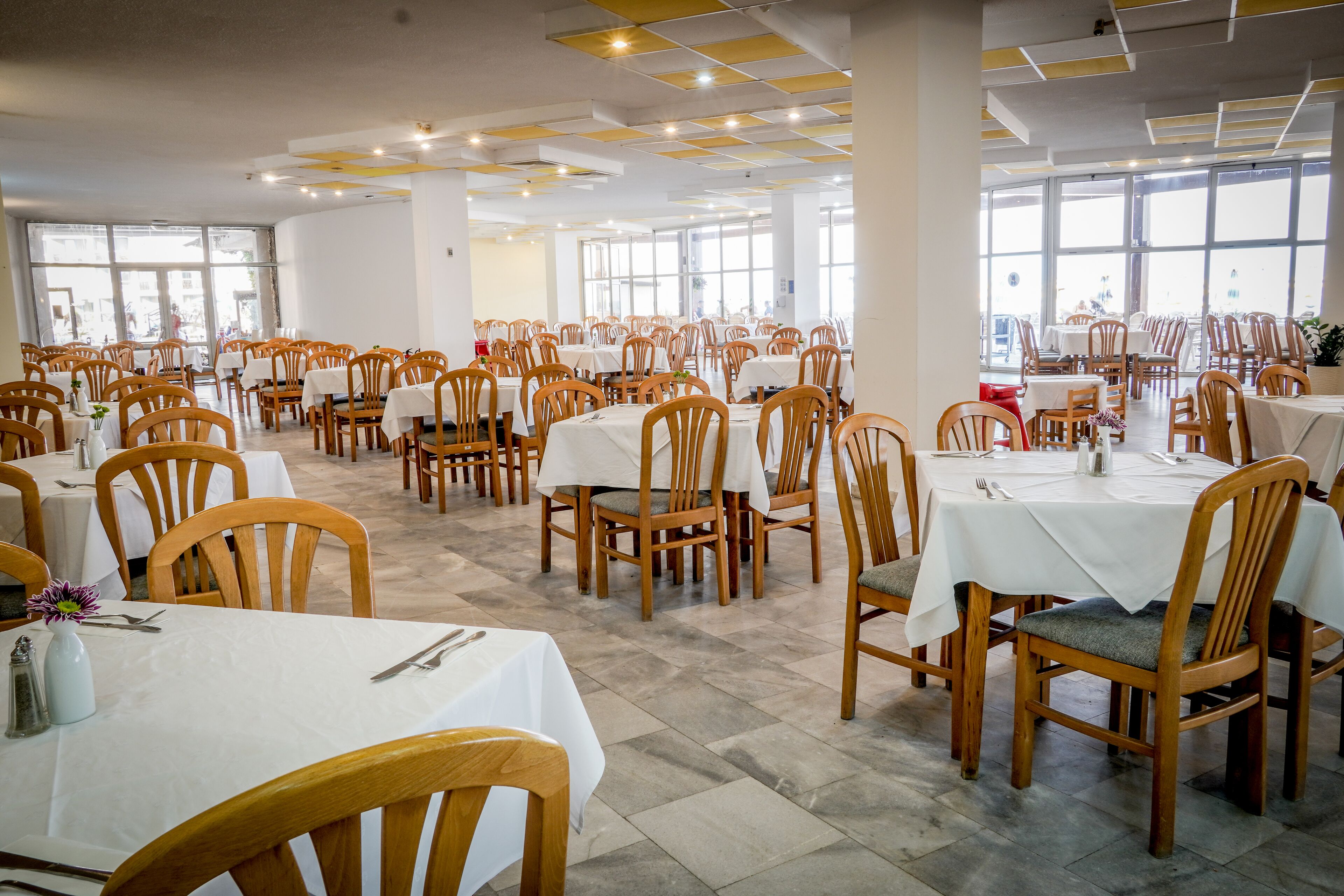A spacious dining area with multiple tables and chairs, large windows, and a tiled floor.