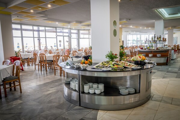 A spacious dining area with round tables and chairs, a buffet counter with various food items, and large windows providing natural light.