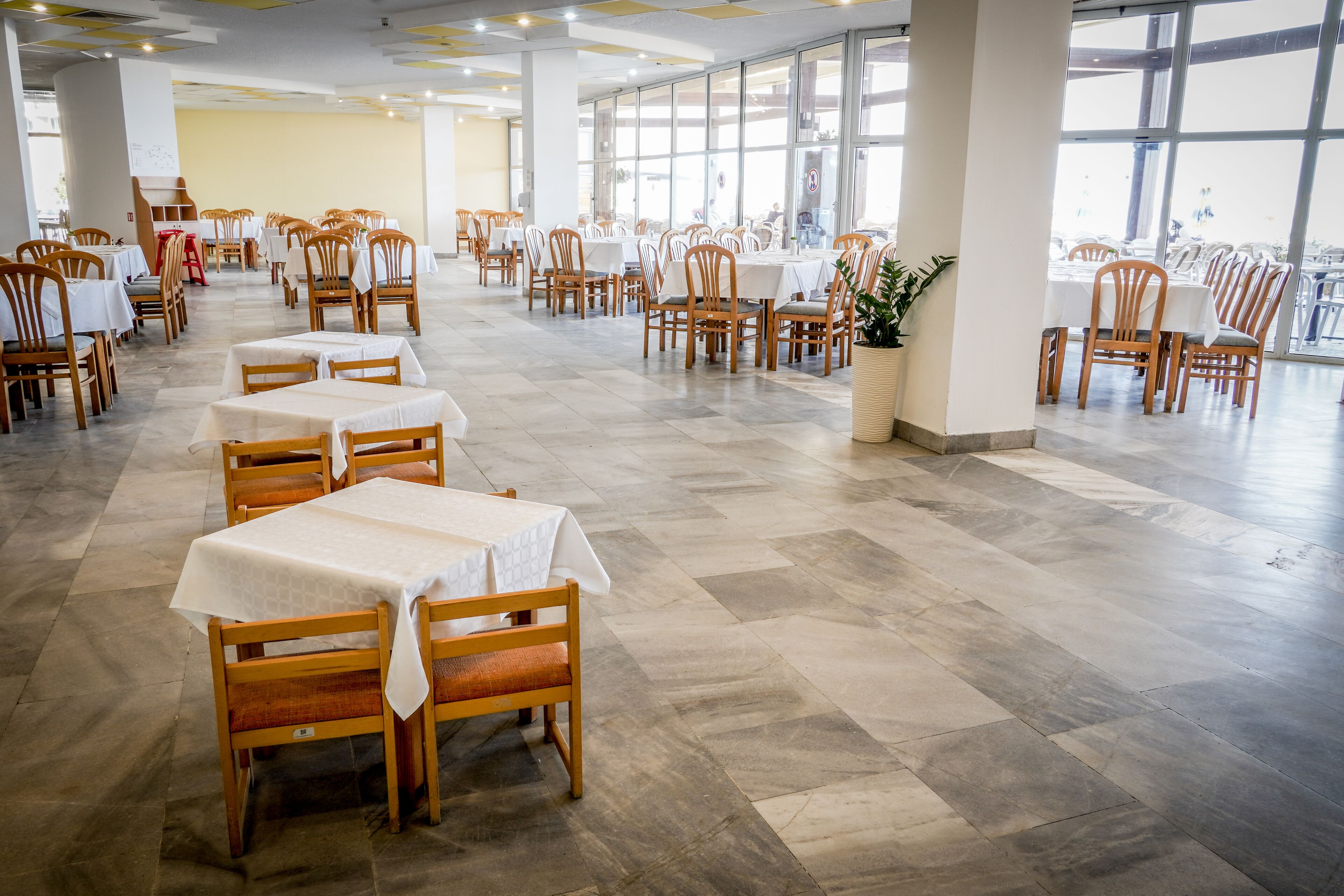 A spacious dining area with multiple tables and chairs, large windows, and a tiled floor.