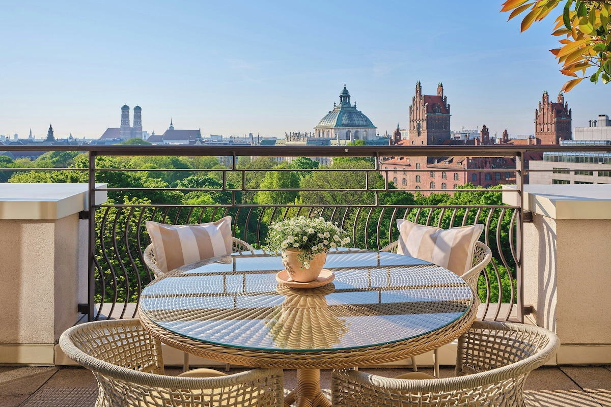 A round glass table with wicker chairs on a balcony overlooking a cityscape with historic buildings.