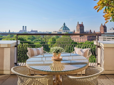 A round glass table with wicker chairs on a balcony overlooking a cityscape with historic buildings.