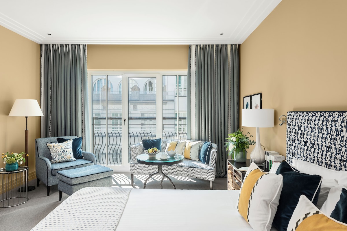 Ein Hotelzimmer mit einem Bett, einem Sofa, einem kleinen runden Tisch mit einer Obstschale und Blick auf ein Gebäude durch große Fenster.