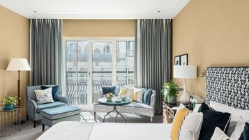 Quarto de hotel com uma cama, um sofá, uma mesinha redonda com uma tigela de frutas e vista para um edifício através de grandes janelas.