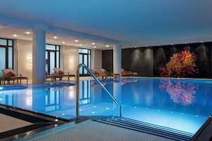 An indoor swimming pool with clear water, a glass barrier, and a seating area with chairs and a table.