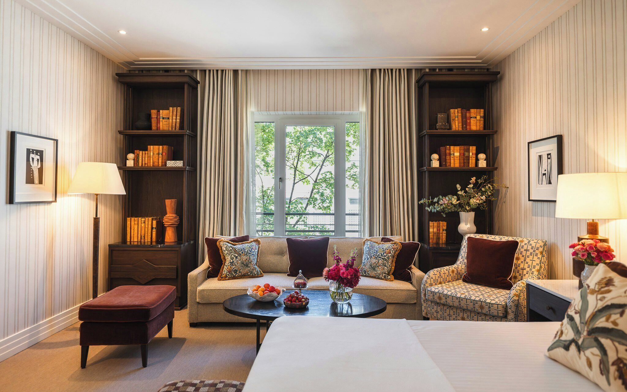 Habitación de hotel con sofá, sillones, una mesita de centro, estantería y ventana con cortinas.