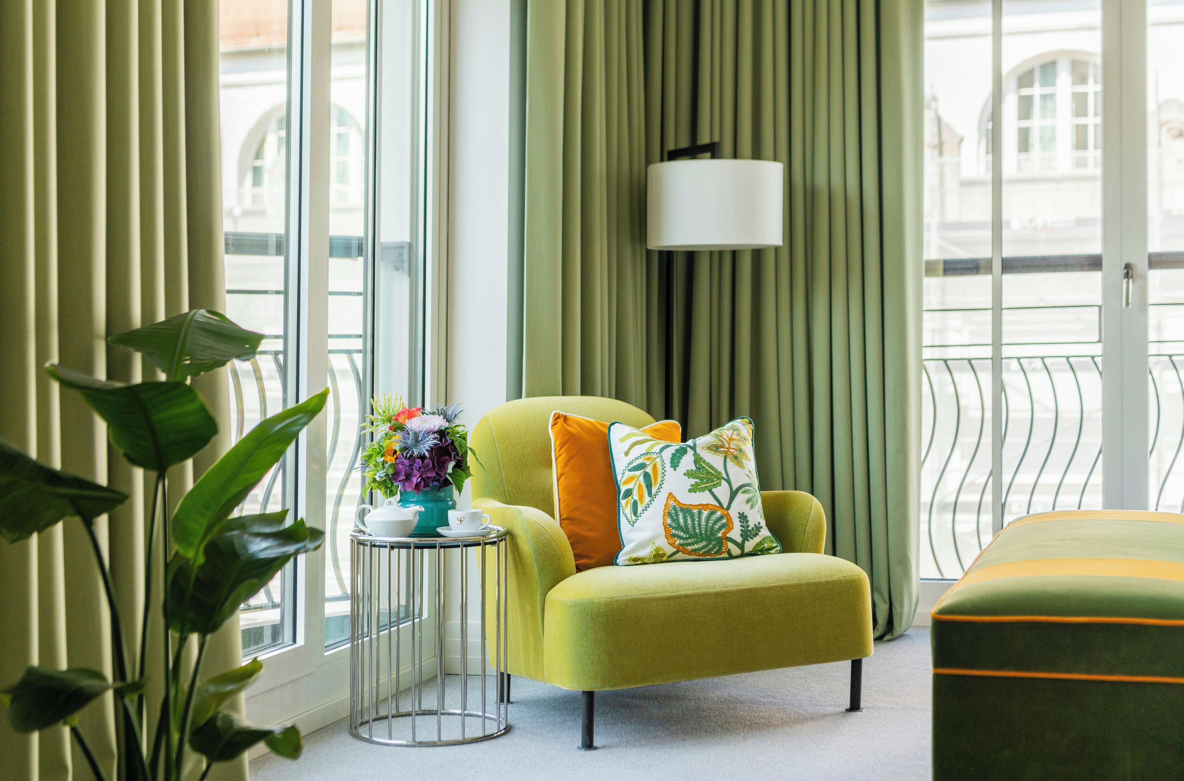 Una sala moderna con un sillón amarillo, una mesita de noche con un jarrón de flores y vistas a la ciudad a través de amplios ventanales.
