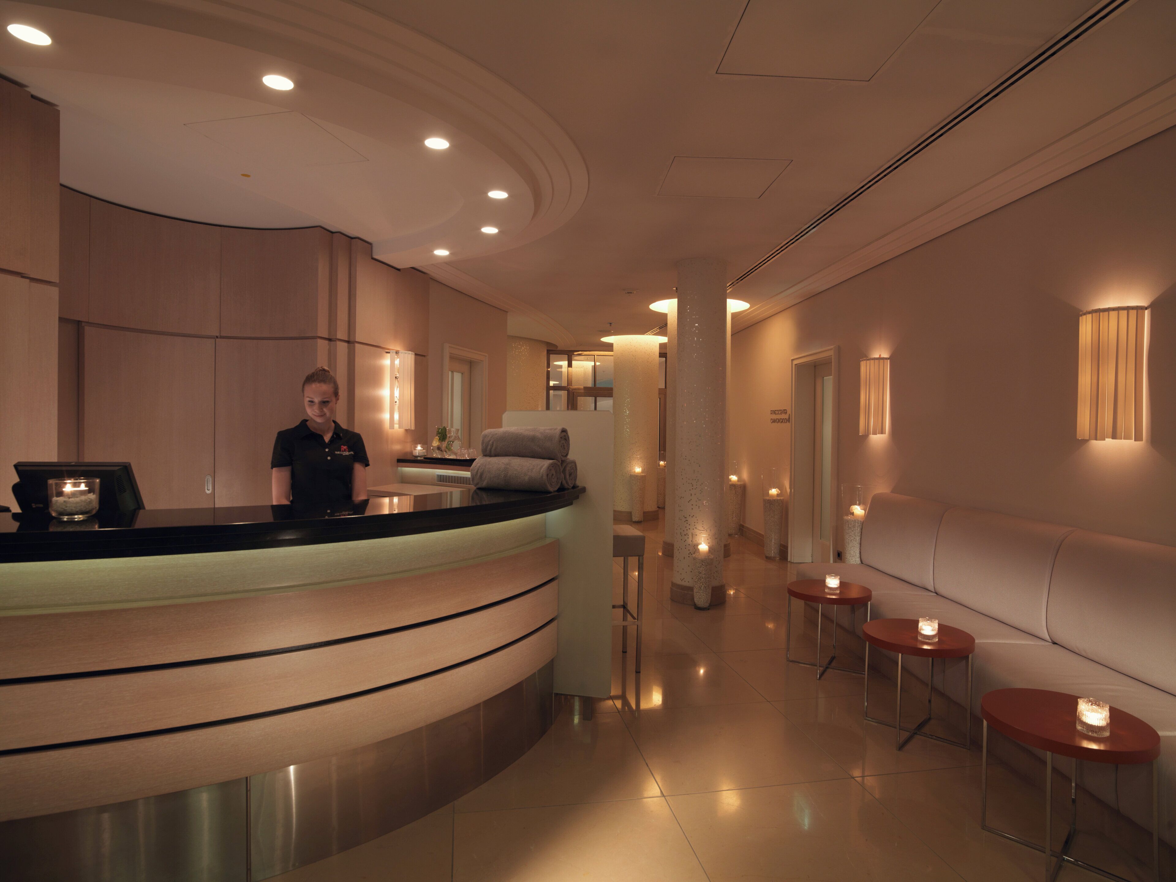 A hotel reception area with a curved counter, a receptionist, and a hallway with seating and wall-mounted lights.