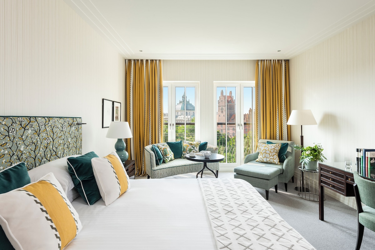Ein Hotelzimmer mit einem großen Bett, zwei Sitzgelegenheiten, einem kleinen Tisch und Blick auf ein Gebäude durch die Fenster.