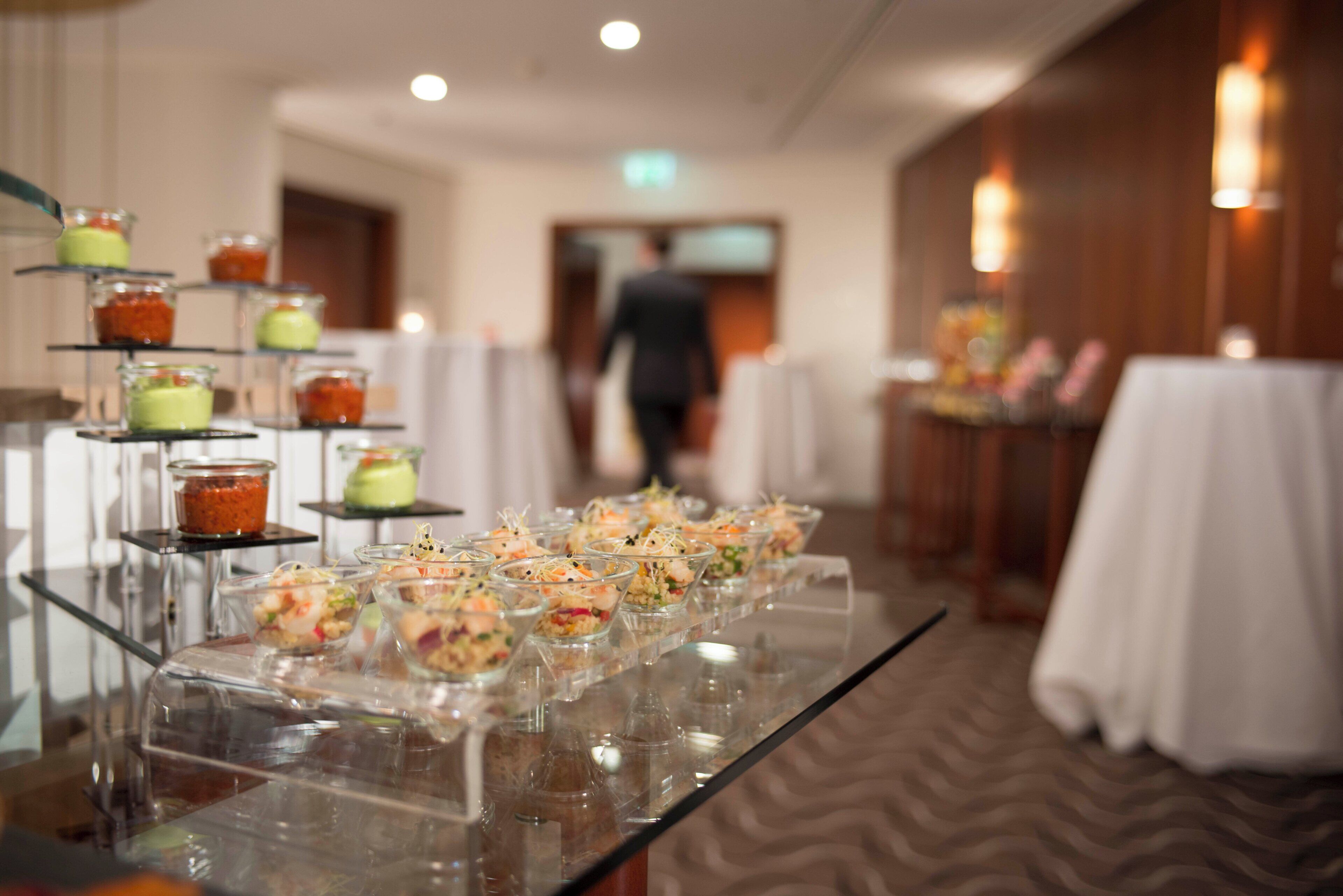 A buffet table with various food items, a person walking in the background, and a carpeted floor.