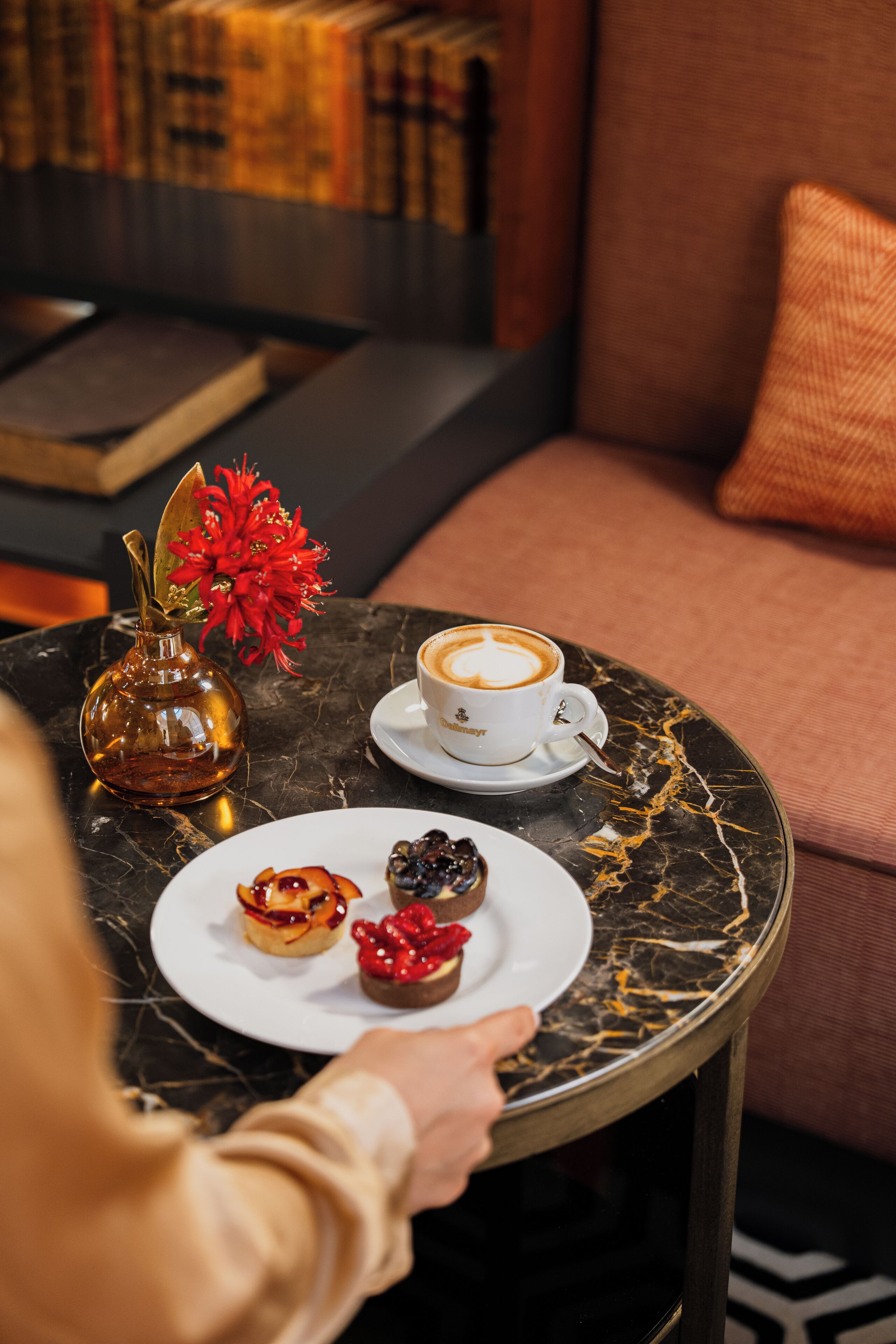 Een tafel met een kop koffie, een kleine vaas met rode bloemen en drie gebakjes.