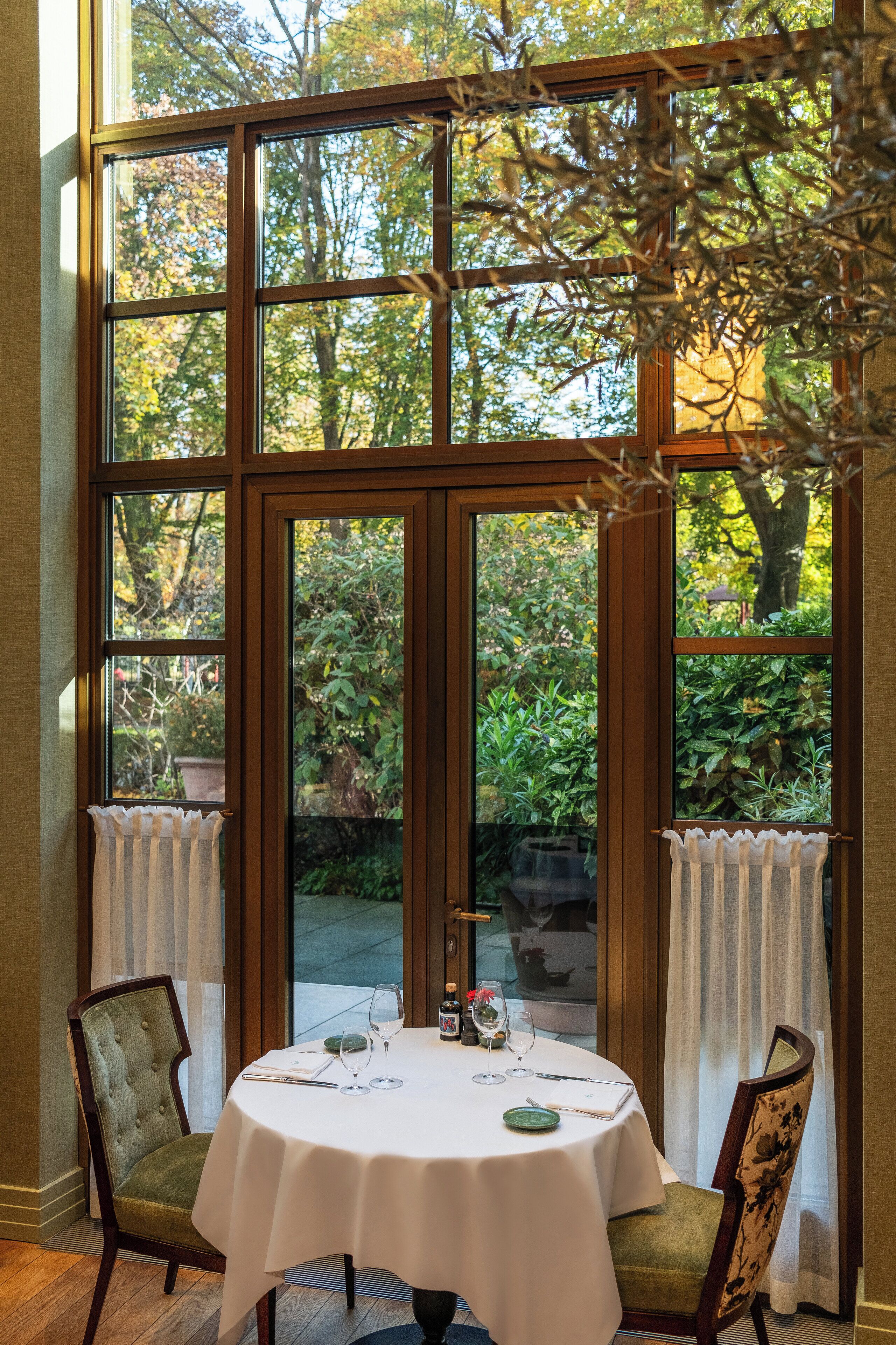 A dining area with a table set for two, large windows, and a view of greenery outside.