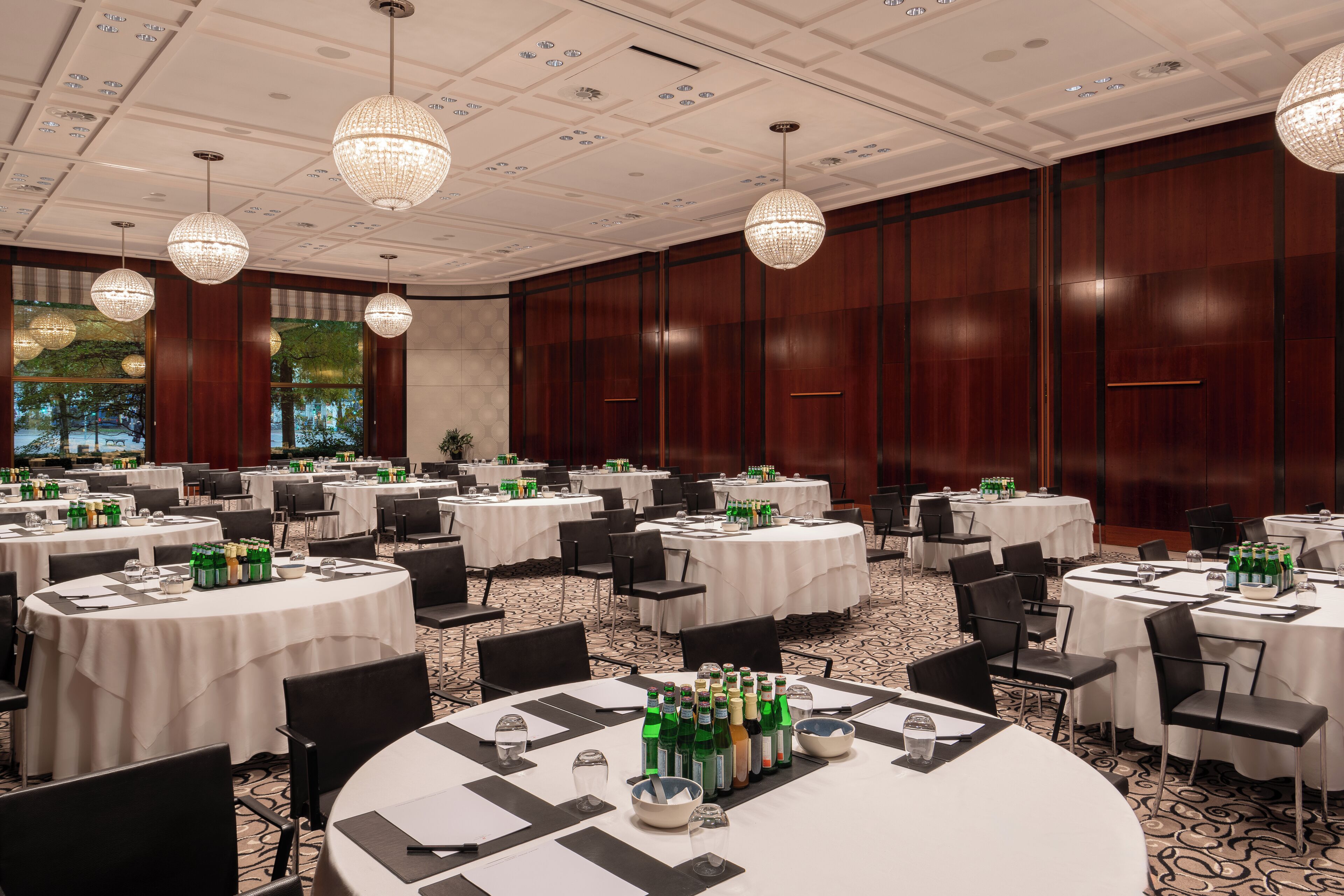 A conference room with round tables, chairs, and a chandelier. Tables are set with water bottles and glasses.