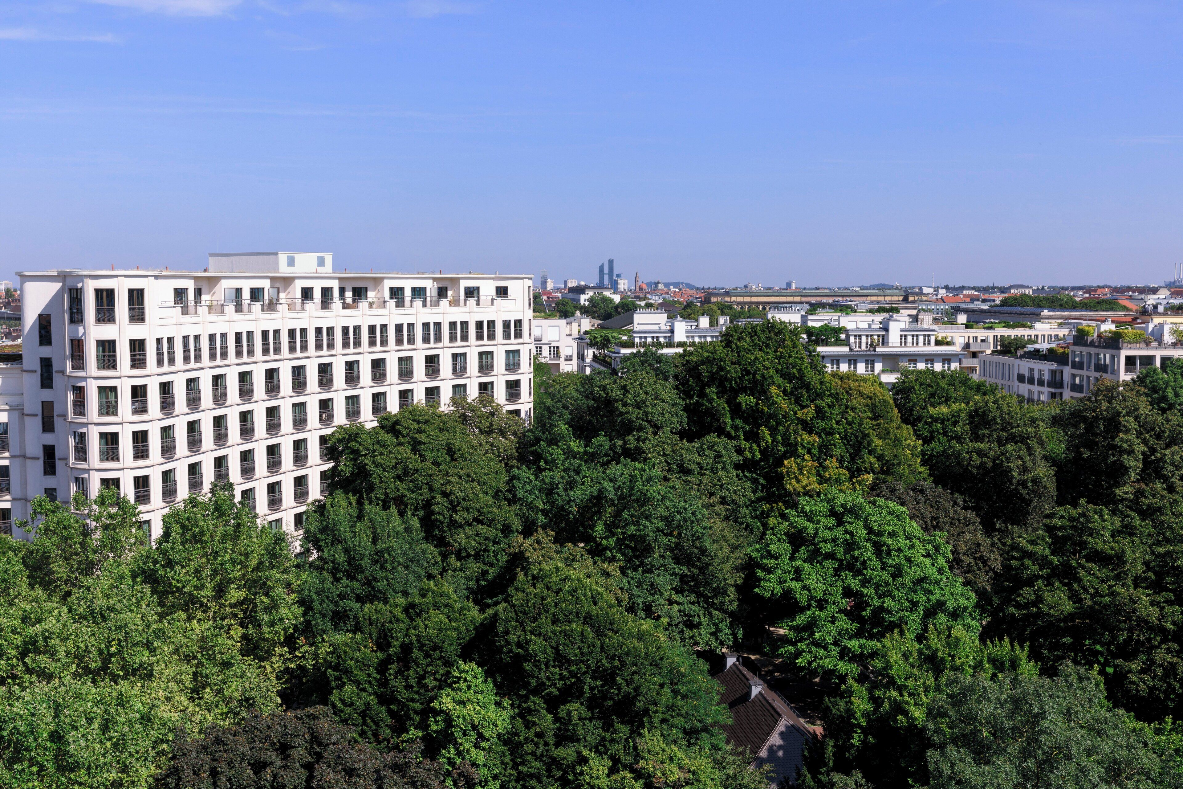 A modern building with a white facade and multiple windows, surrounded by dense green trees.
