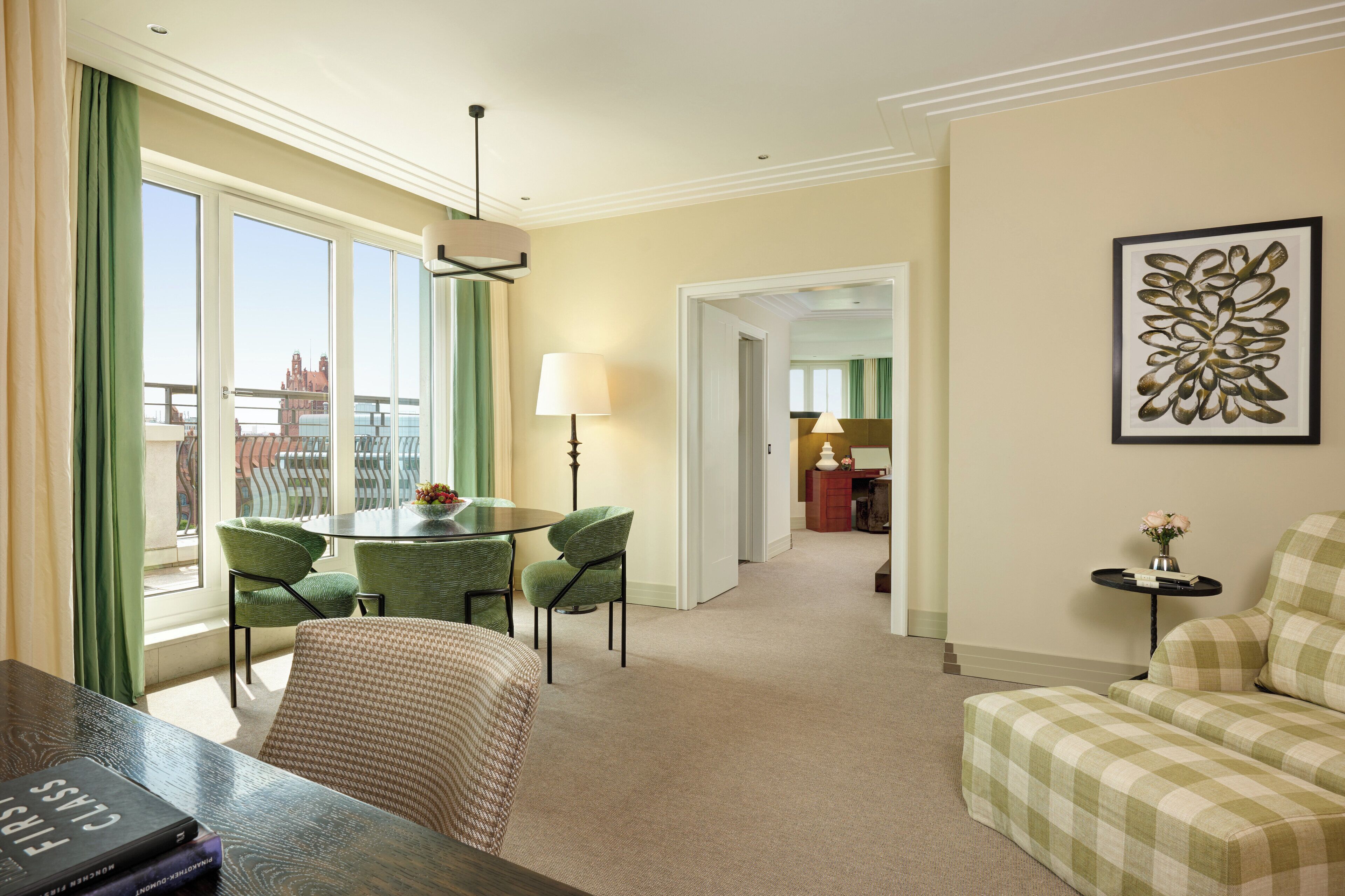 A hotel room with a dining area, a sofa, a table with a lamp, and a framed artwork on the wall.