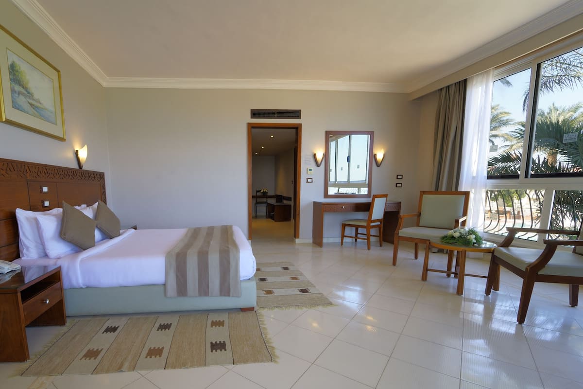 Hotelkamer met een groot bed, een houten nachtkastje, een bureau met stoel en uitzicht op palmbomen door het raam.